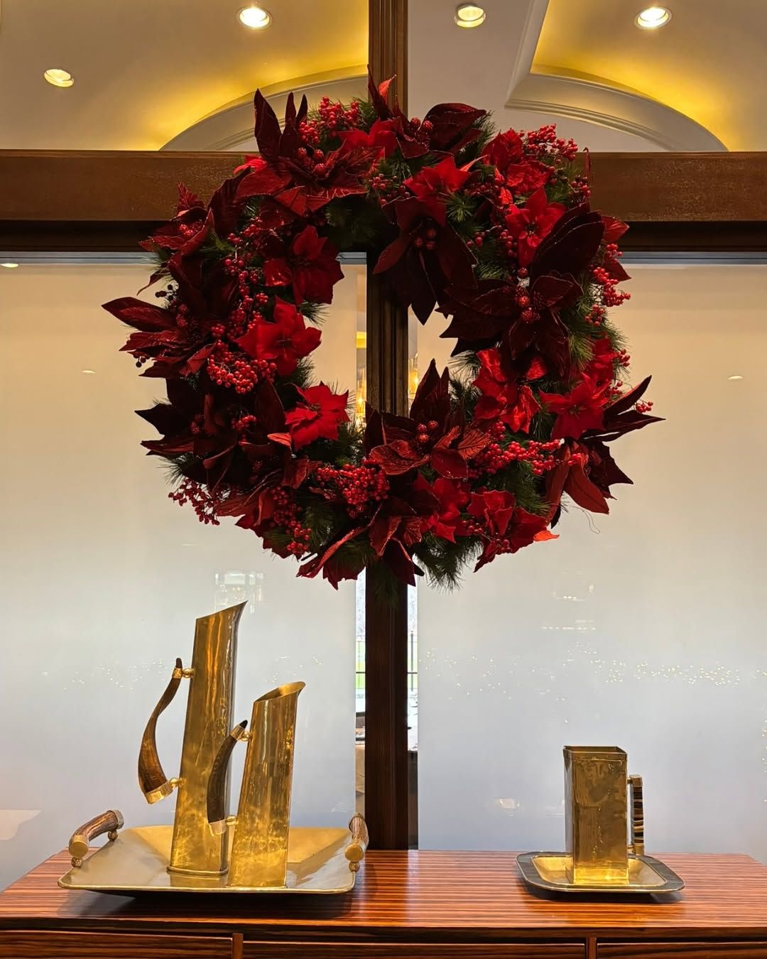 Large wreath of poinsettias hung on windows above a credenza with gold drinkware on top.  