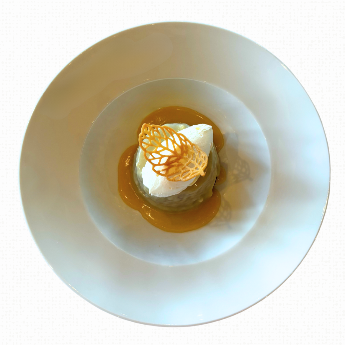 A white bowl of sticky toffee pudding with toffee sauce, chantilly cream, and honey tuile on a white tablecloth-covered table. 