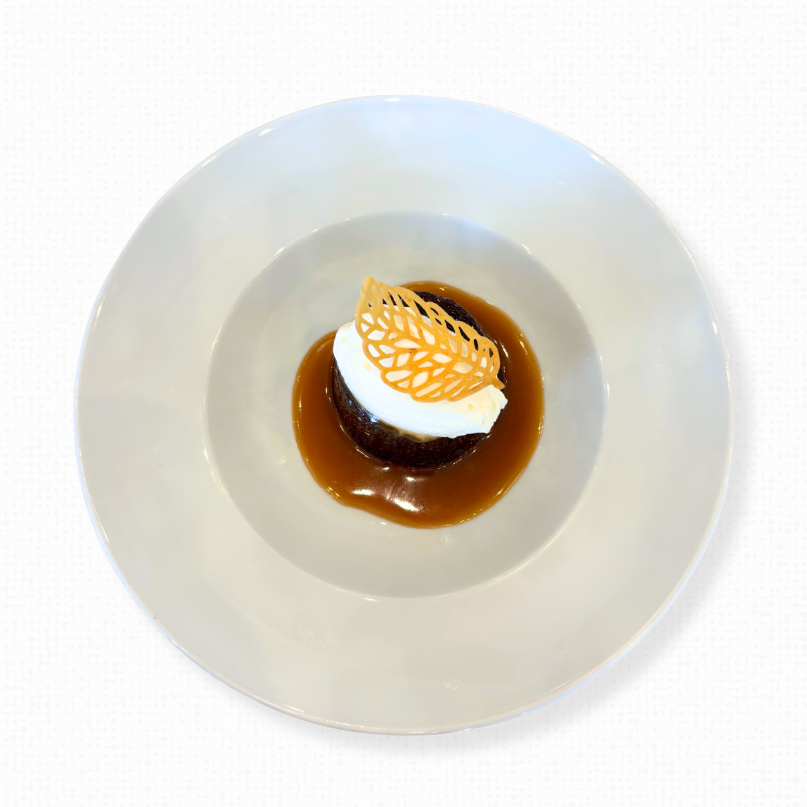A white bowl of sticky toffee pudding with toffee sauce, chantilly cream, and honey tuile on a white tablecloth-covered table. 