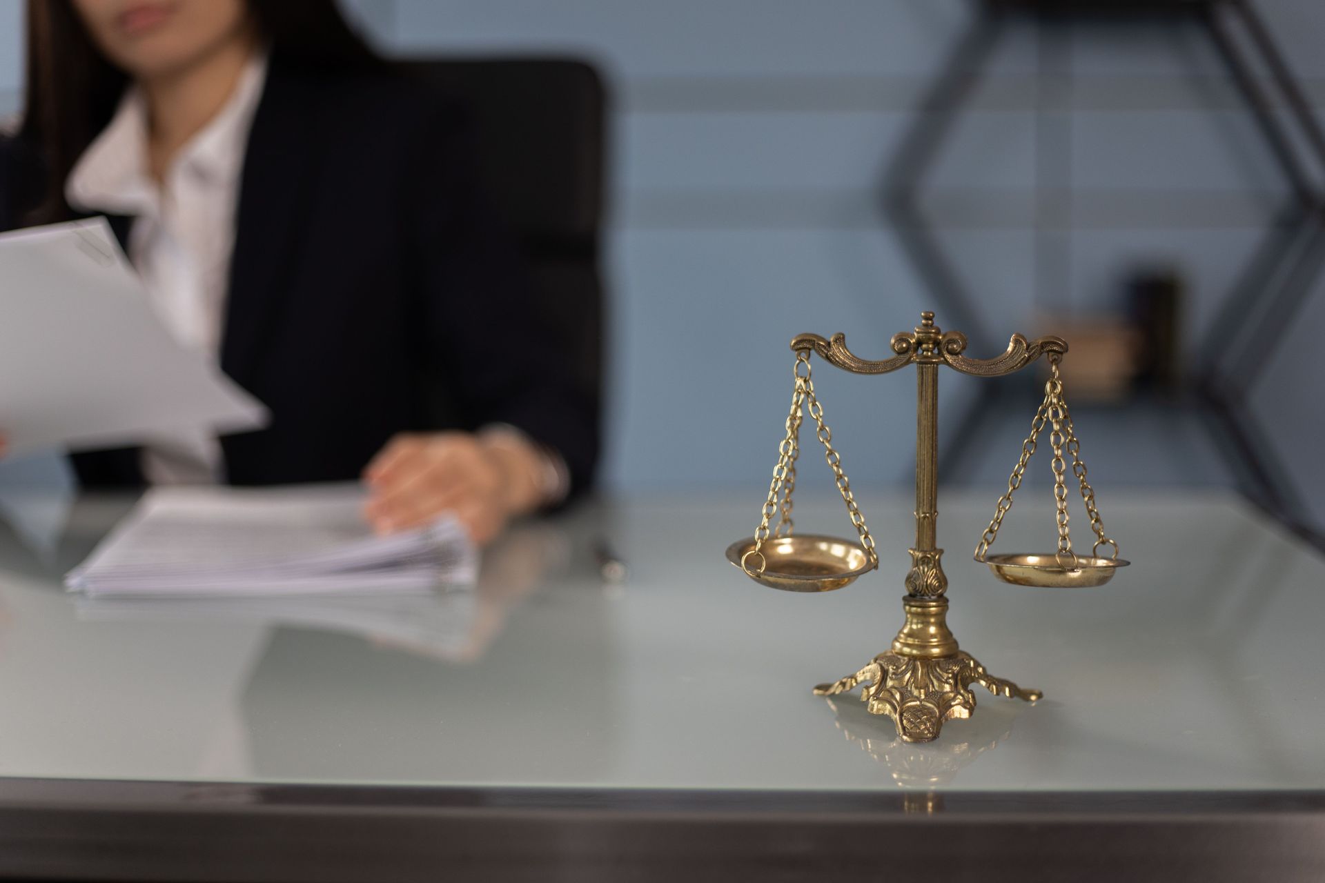 Gold scales of justice on a desk with a blurred person reviewing documents in the background