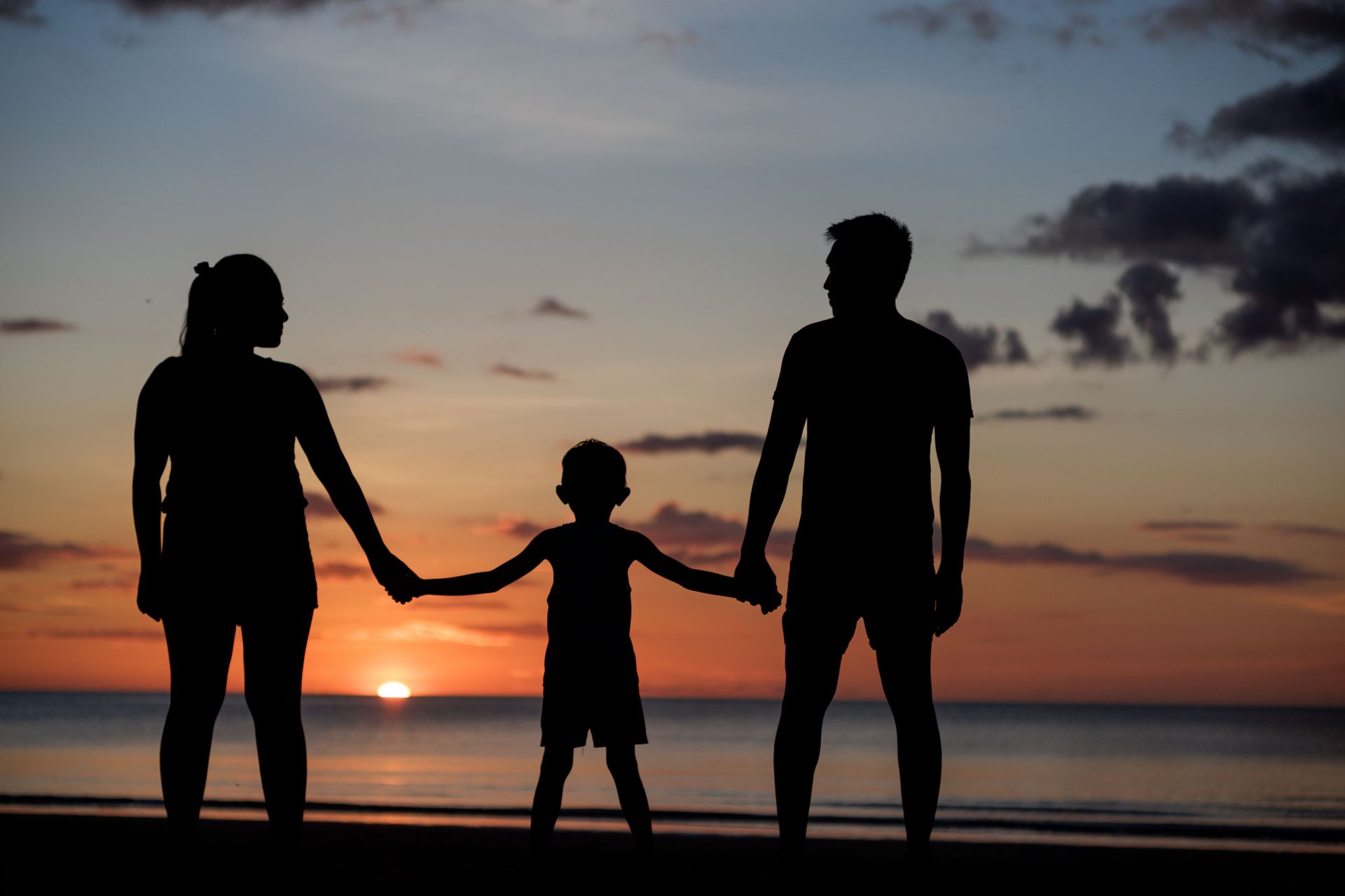 Silhouetted family holding hands on a beach at sunset with ocean waves and glowing sky.