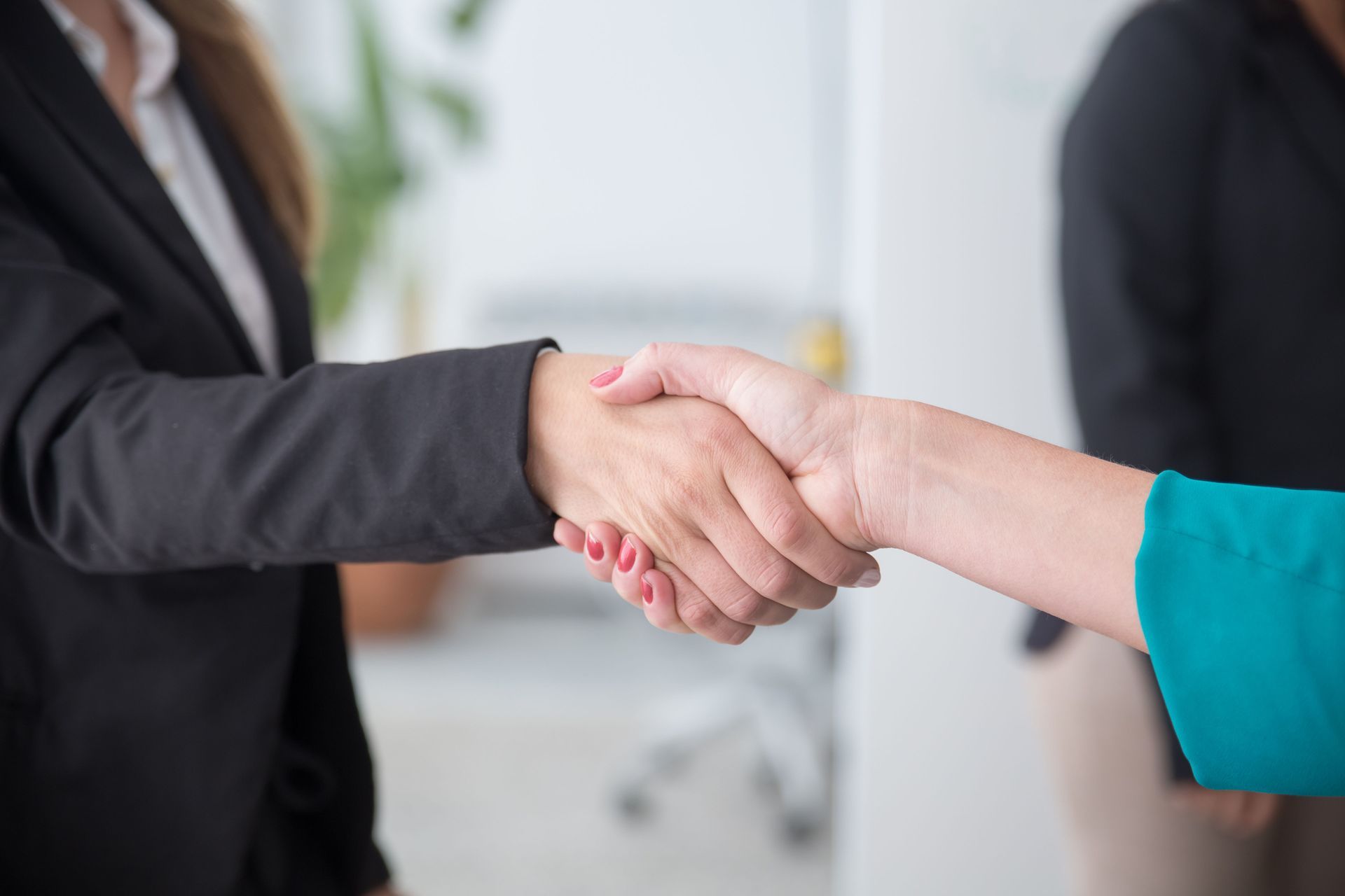Businesspeople shaking hands in an office, one in a black suit and one in a teal sleeve.