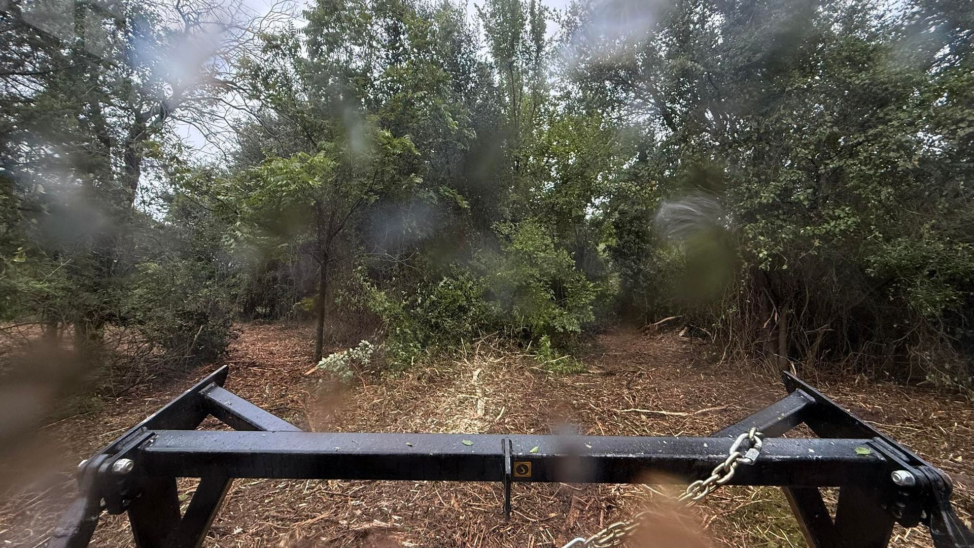 View from a tractor cab, approaching trees on a muddy path, rain on windshield, dark metal implement in foreground.