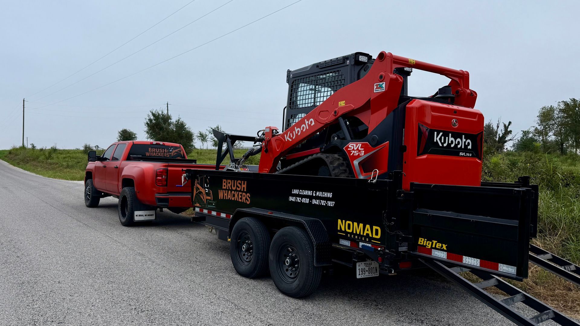 Red pickup truck towing a black trailer with a red Kubota skid steer on a gravel road.