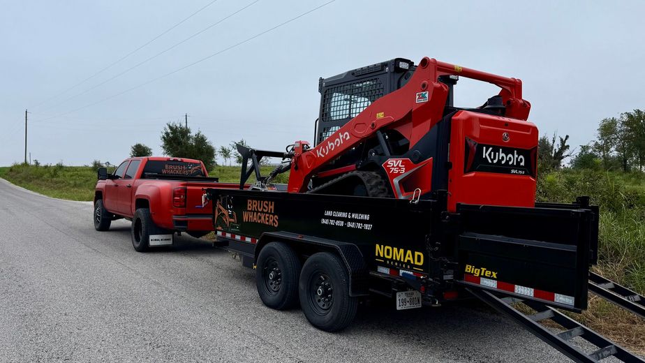 Red pickup truck towing a black trailer with a red Kubota skid steer on a gravel road.