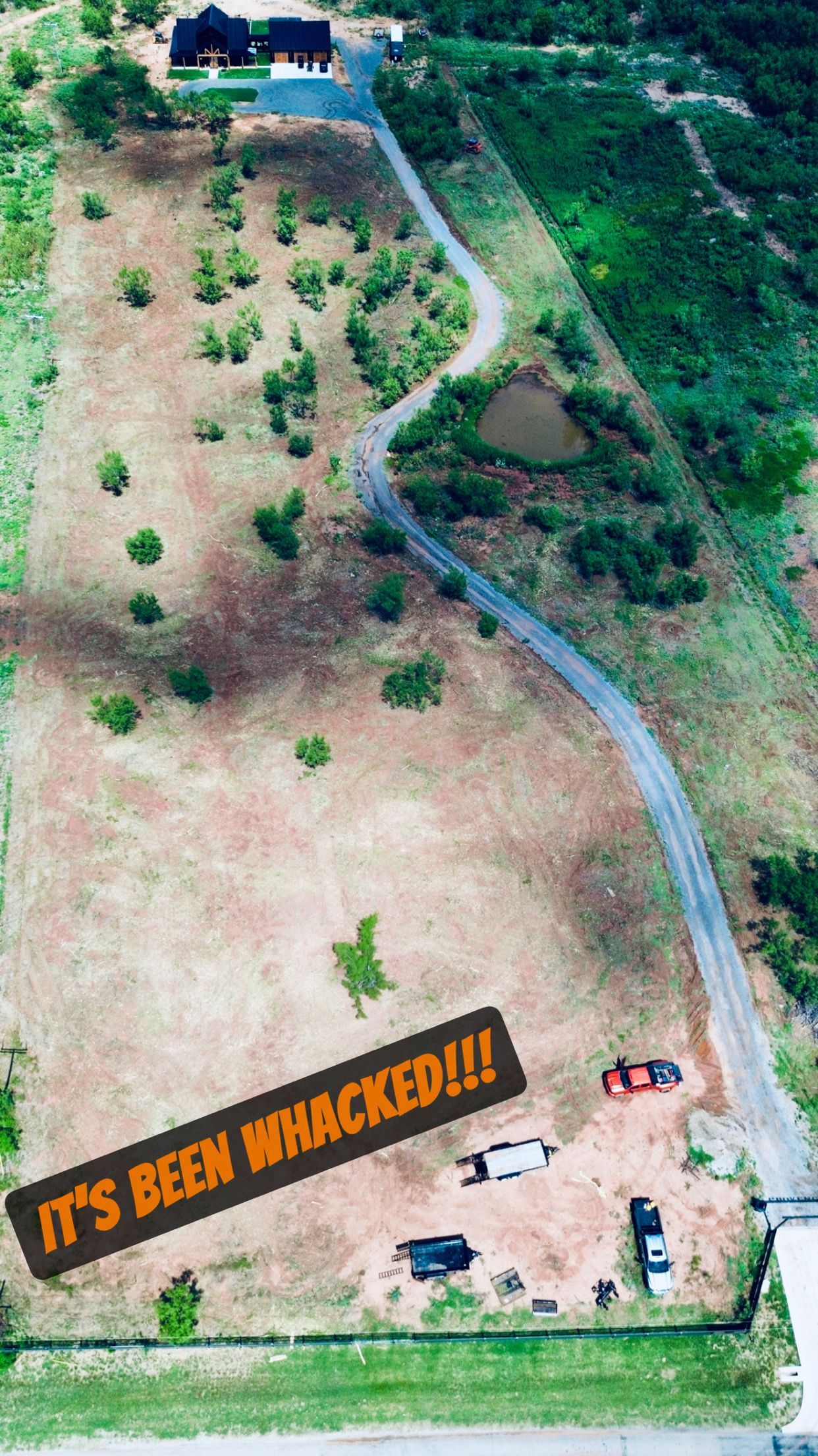 Aerial view of a cleared field with a winding dirt road and a few vehicles parked at the bottom; a house is at the top.