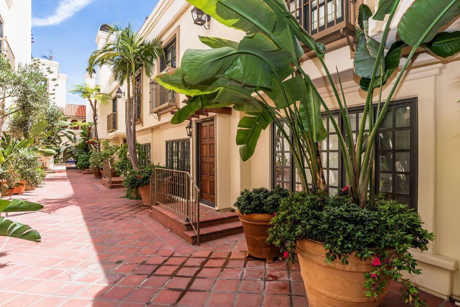Courtyard with terracotta tiles, potted plants, and light-colored buildings with windows and a wooden door.