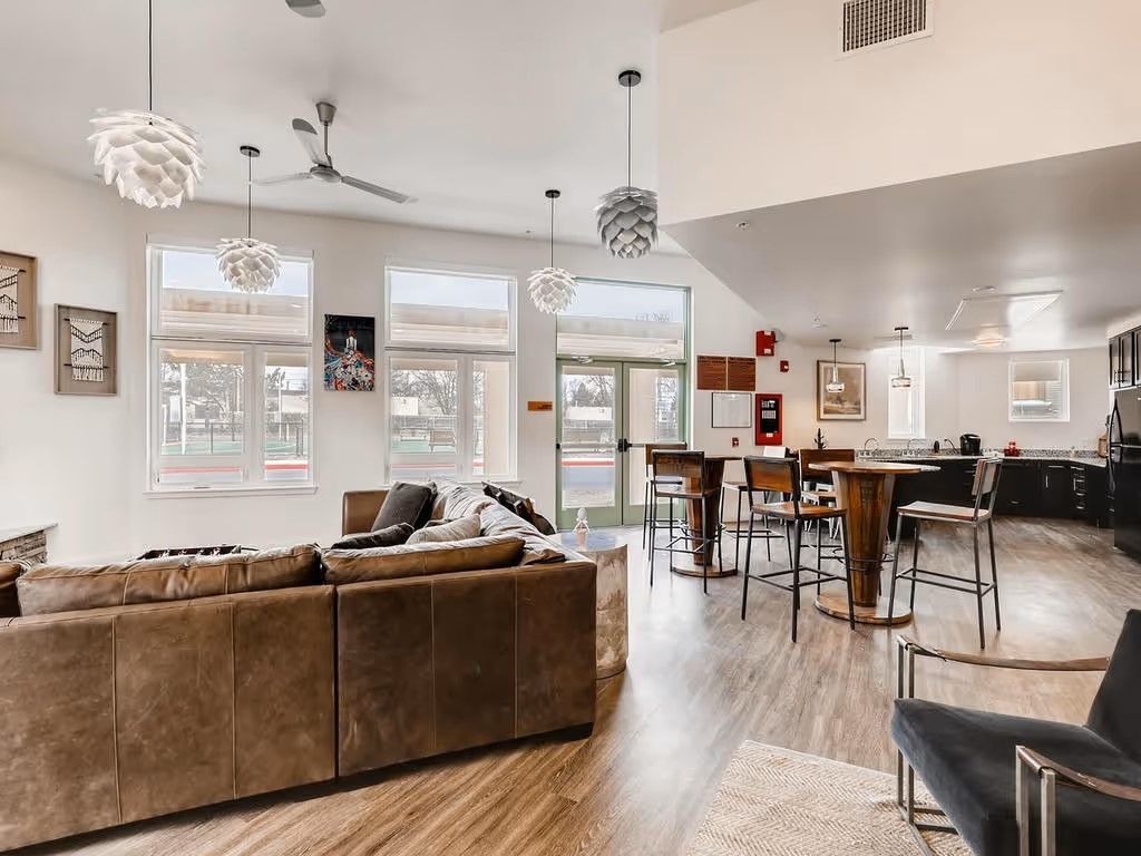 A modern open-plan living room with a brown sofa, high-top dining area, and large windows with natural light.