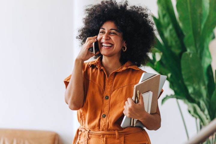 A smiling person in a rust-colored outfit talks on a smartphone while holding notebooks and a laptop.