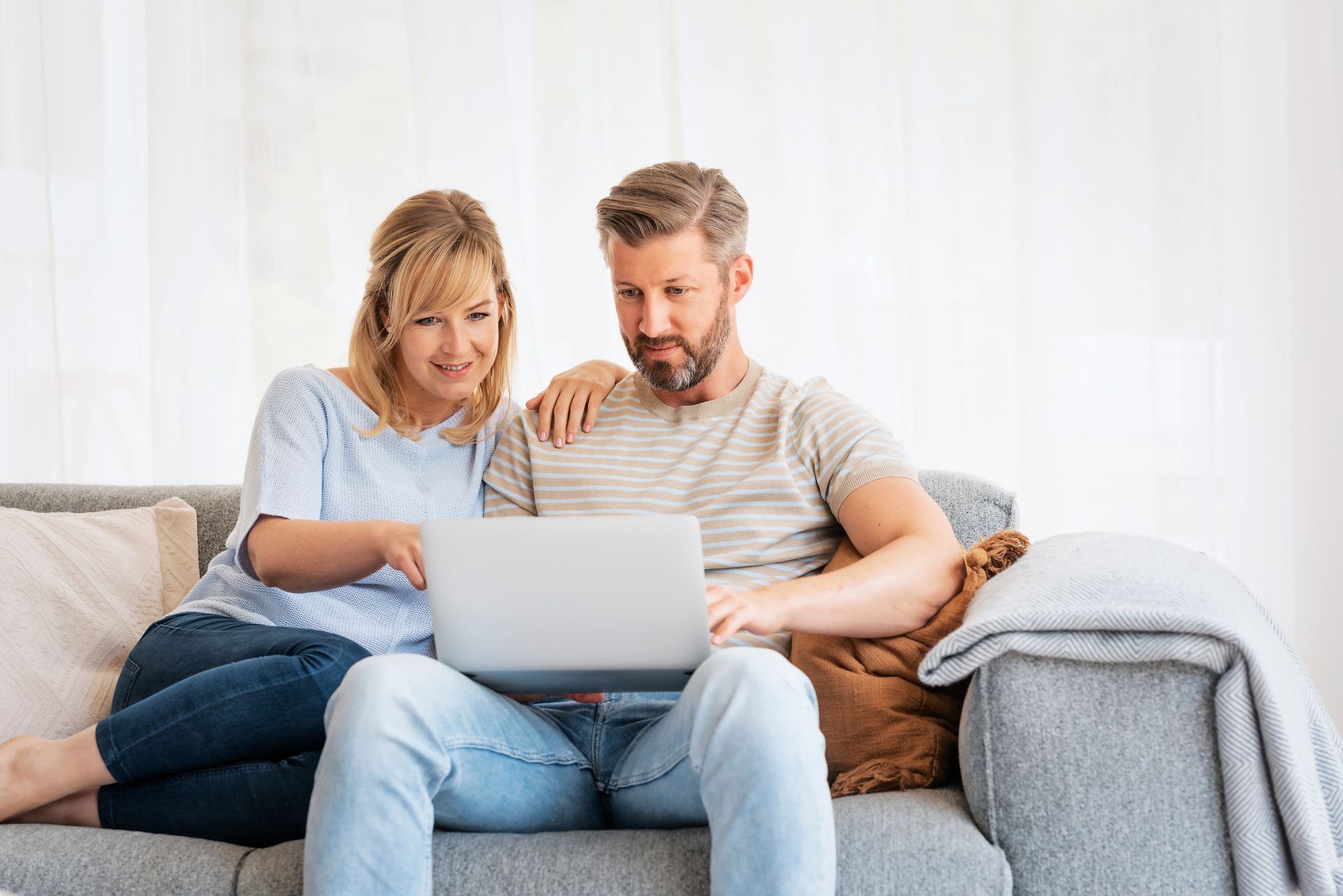 A person pointing at a laptop screen while sitting next to another person on a gray couch in a brightly lit room.