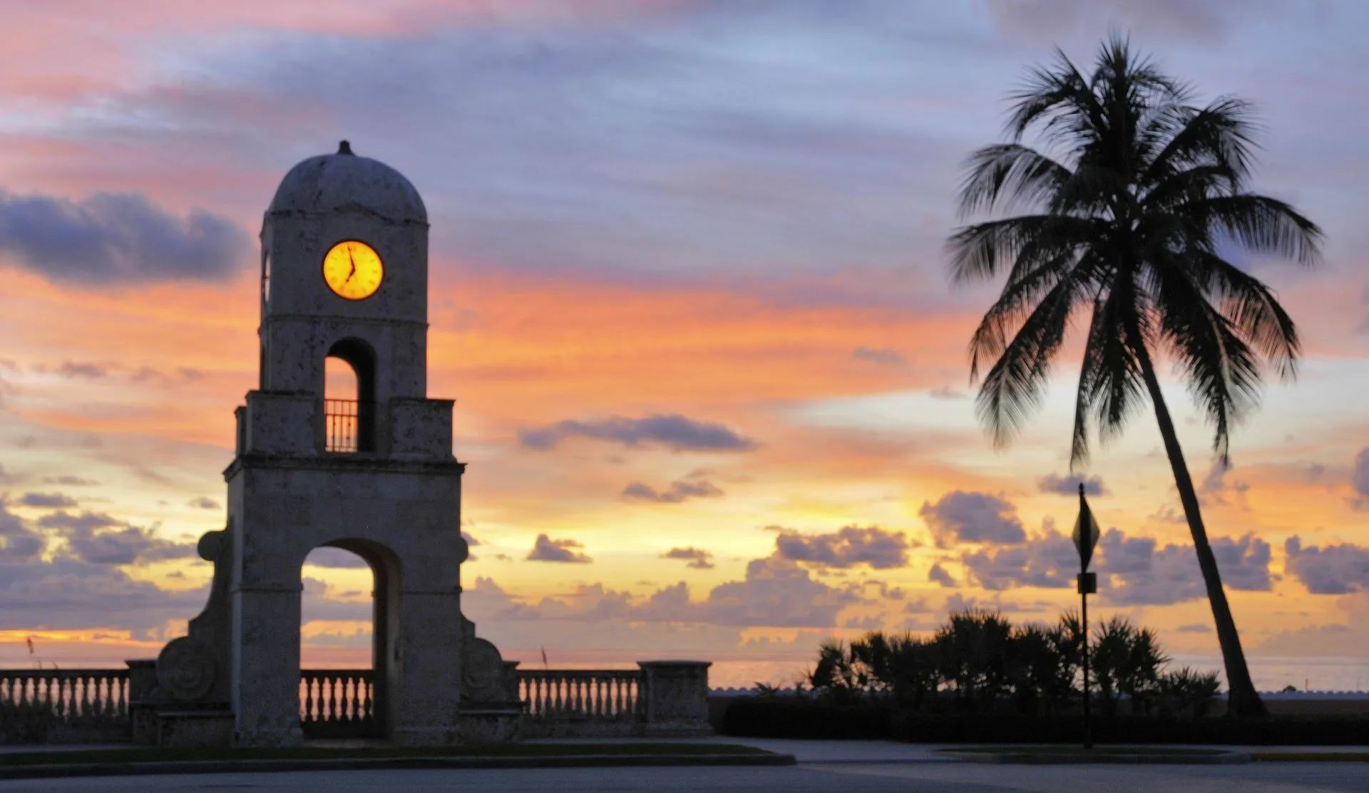 Clock tower by the ocean at sunset with a palm tree silhouette and colorful sky.