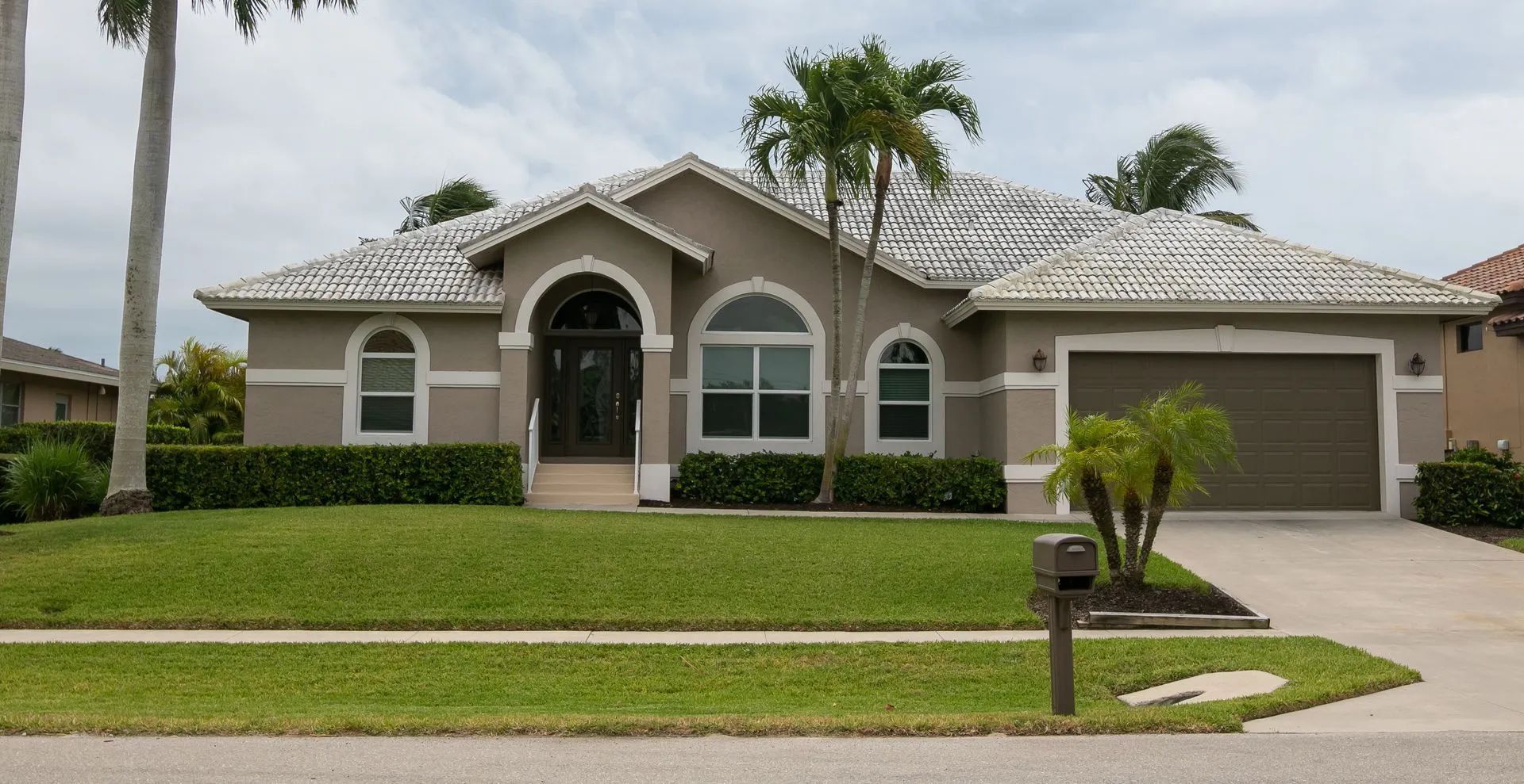 Tan suburban house with green lawn, palm trees, and a two-car garage.