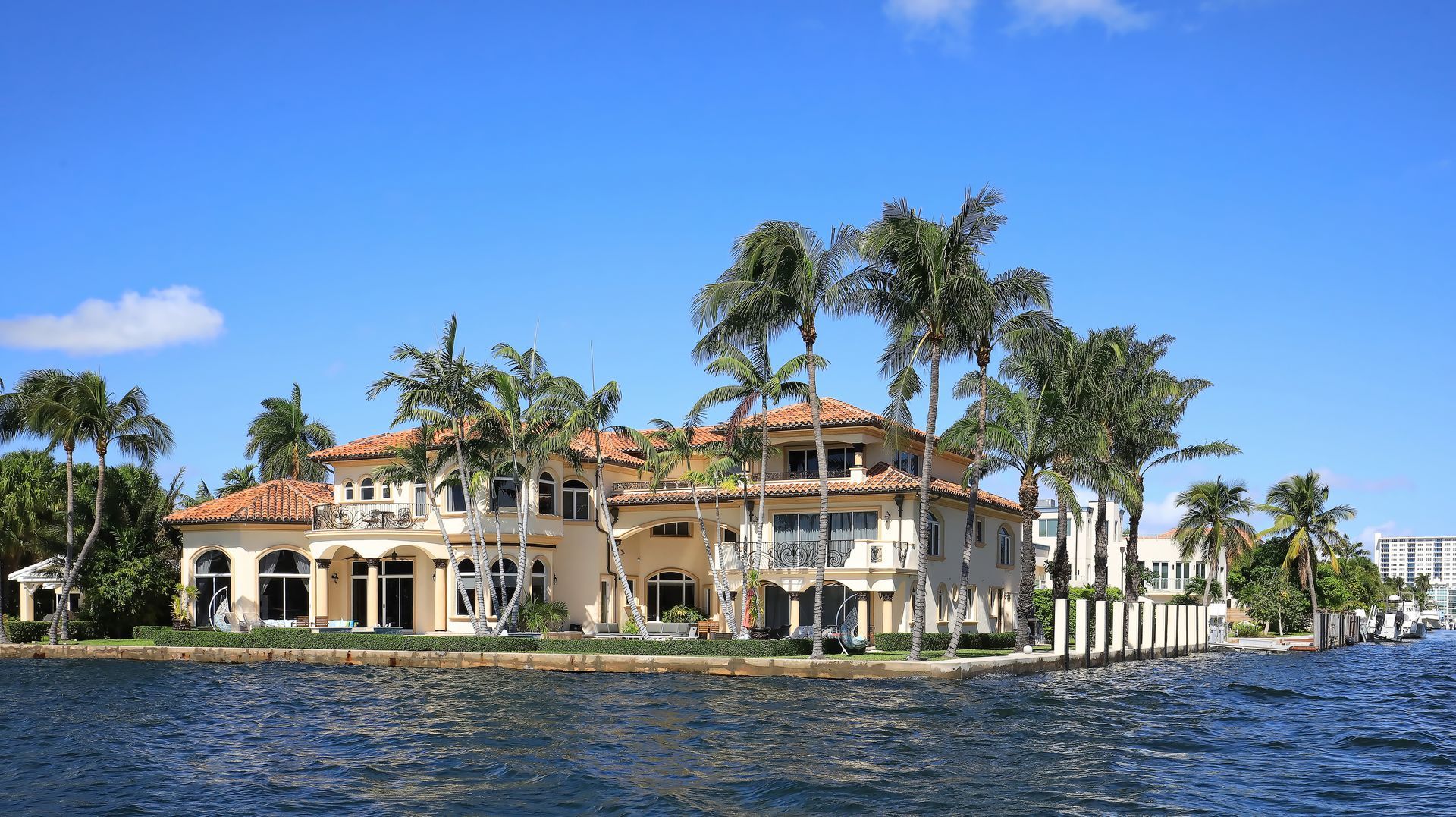 Large beige waterfront mansion with palm trees, blue sky, and water.