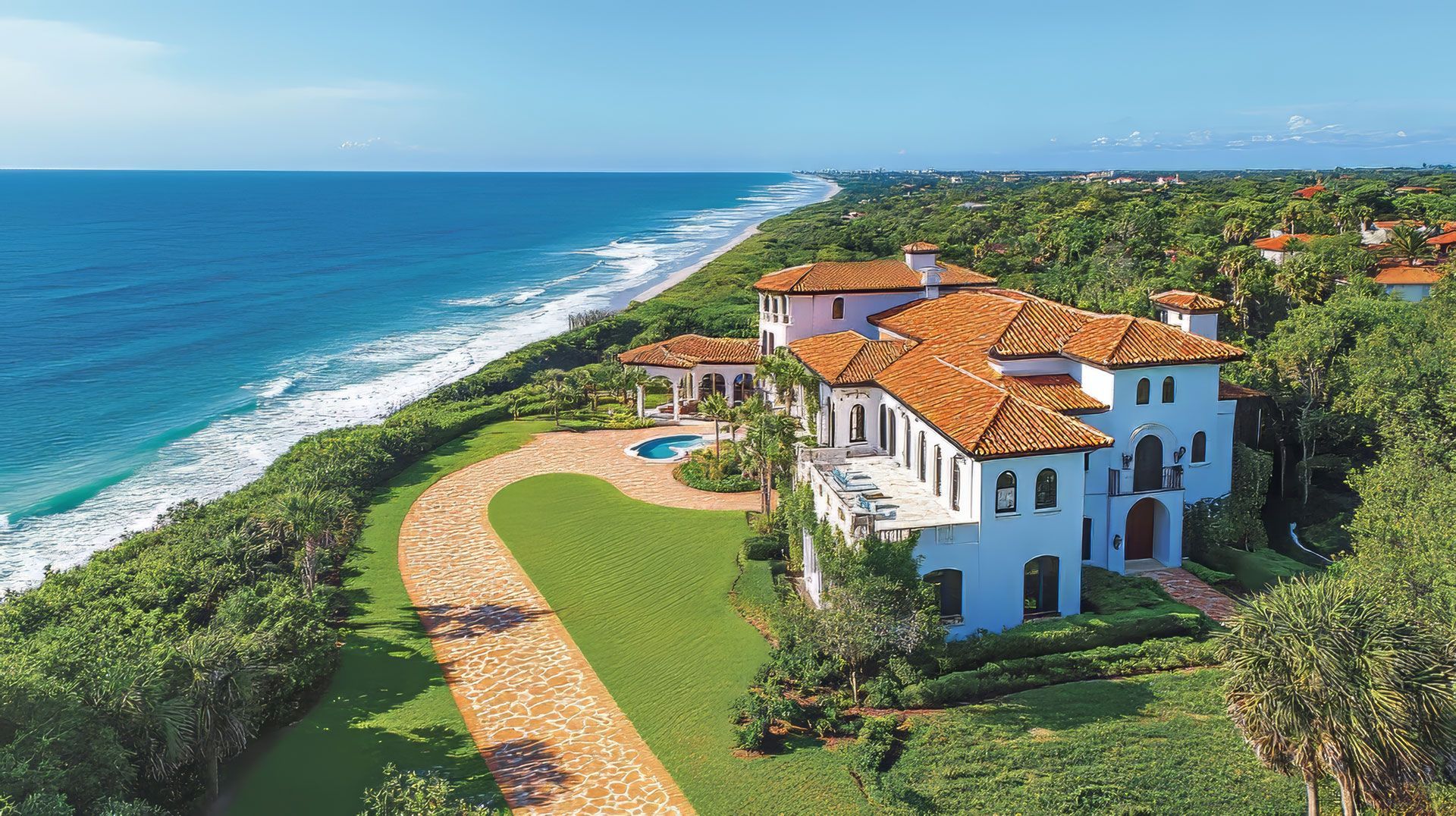 Coastal mansion with terracotta roof, ocean view, green lawn, and winding driveway.