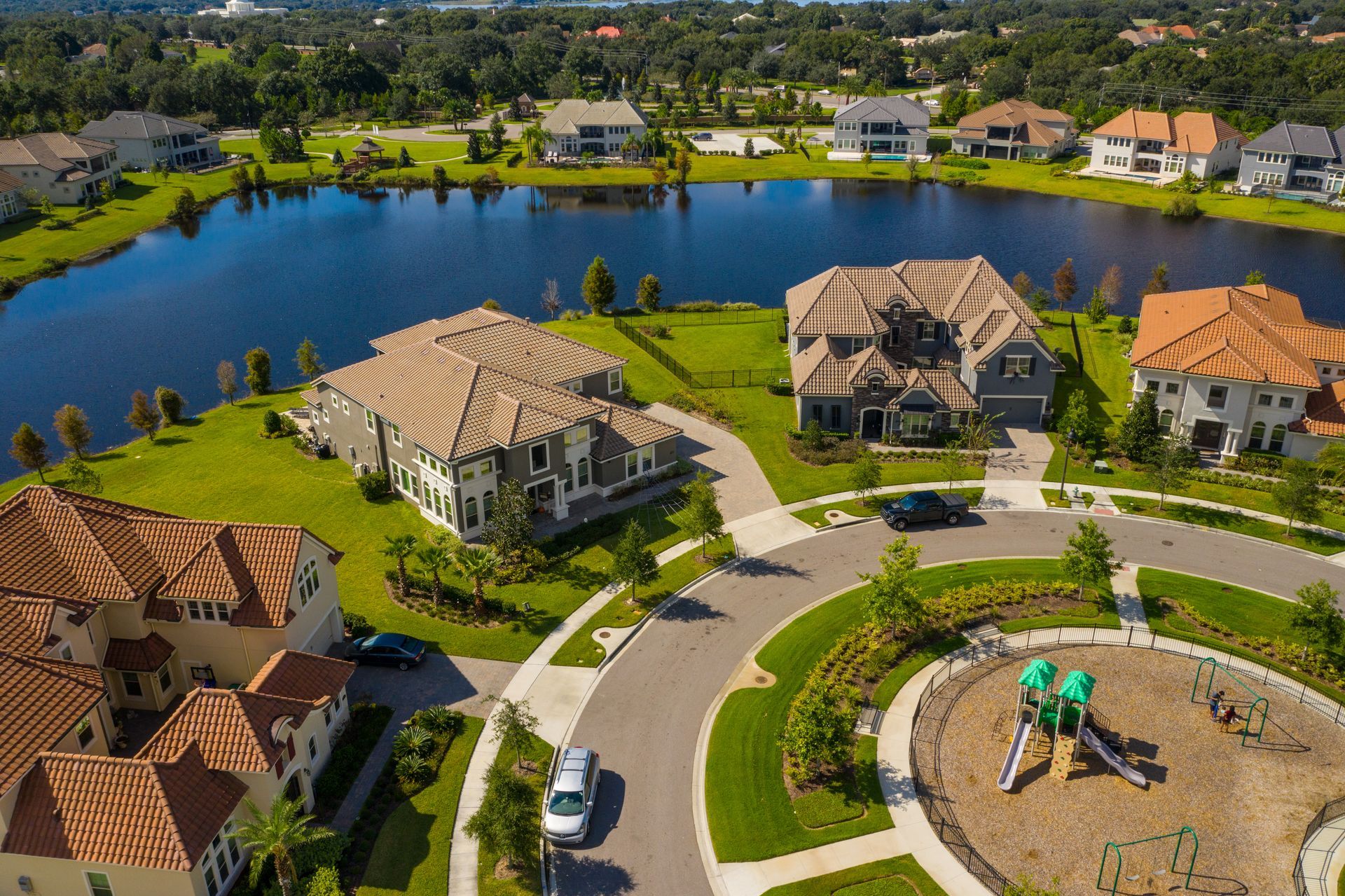 Houses near a lake, some with clay tile roofs, circling a small playground.
