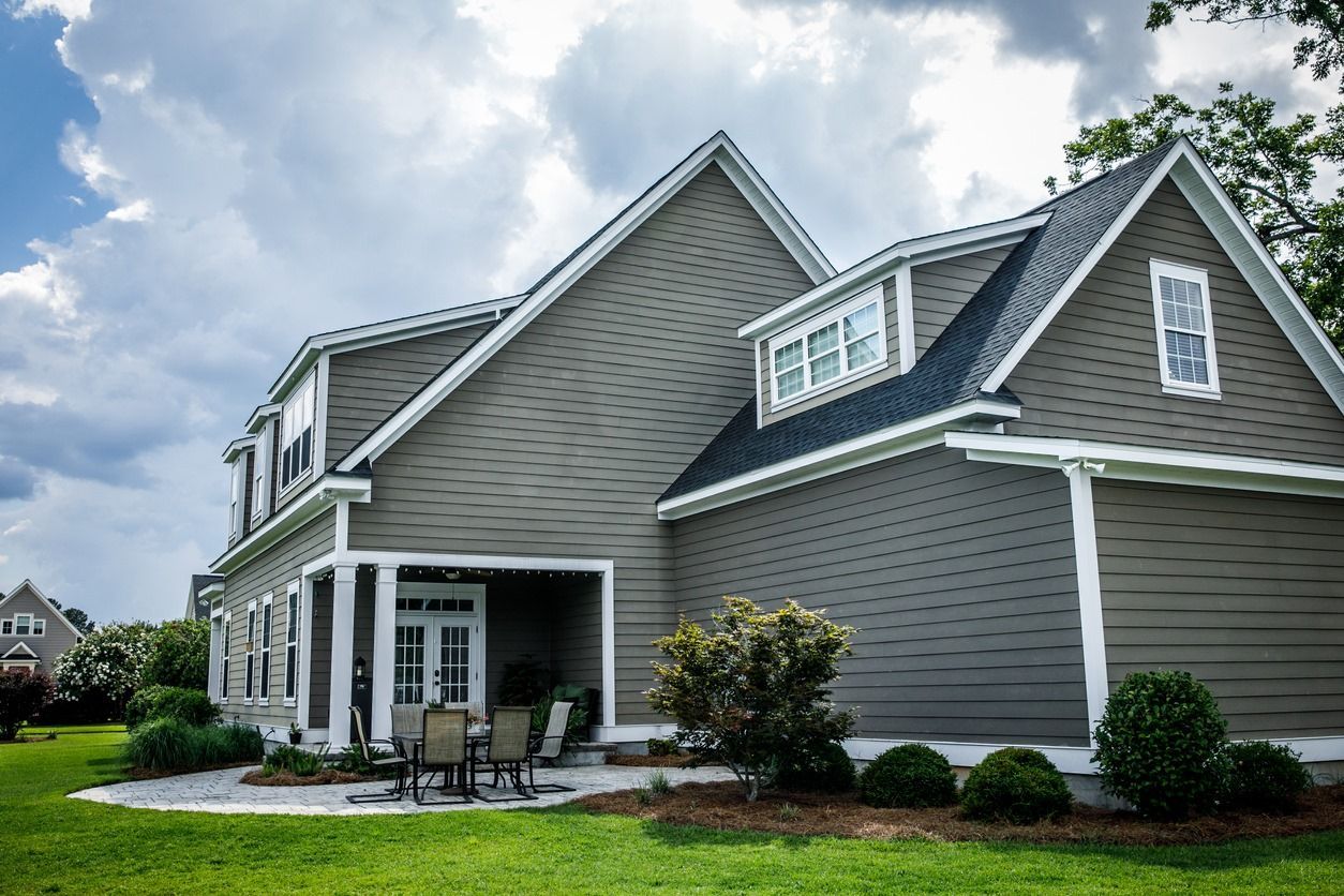 Gray house with white trim and a small patio area. Green lawn and a cloudy sky.