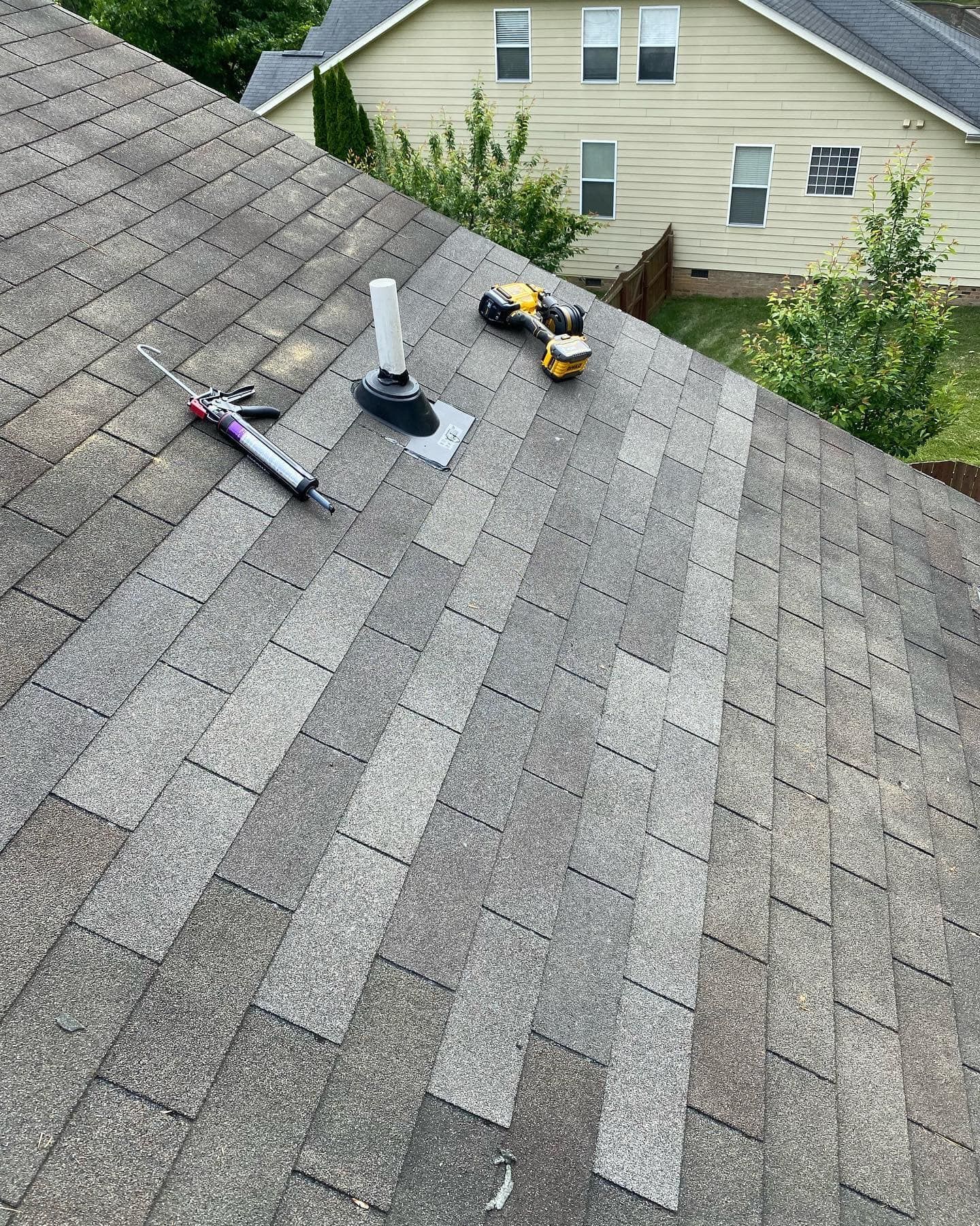 A rooftop with shingles, caulk gun, drill, and vent pipe; sunlight. Houses and trees in the background.