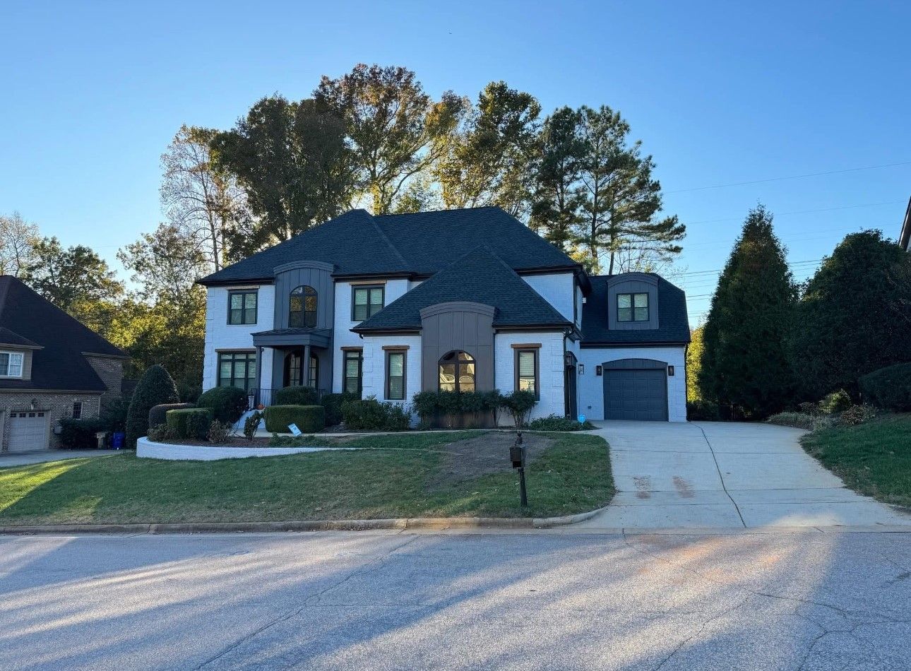Two-story white house with gray accents and a dark roof, set on a slight hill with a driveway, trees, and blue sky.