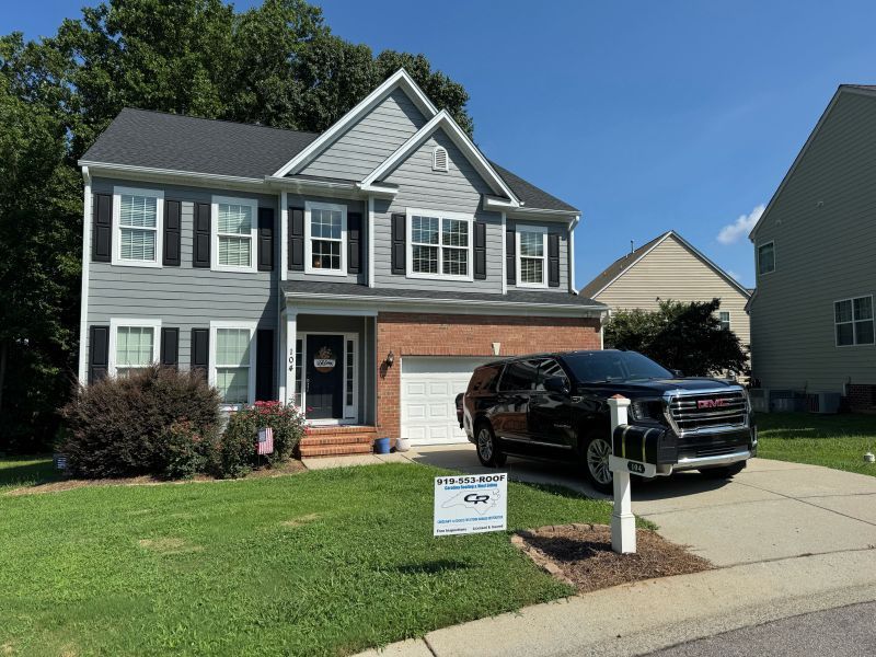 Two-story house with gray siding, black shutters, and a black SUV parked in the driveway. A sign on the lawn shows a business name.