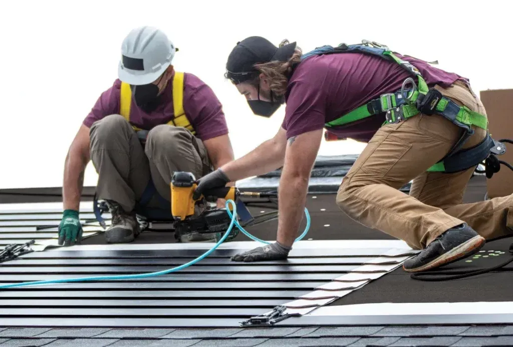 Two workers installing solar panels on a rooftop, wearing safety gear and masks. One uses a nail gun while the other assists.