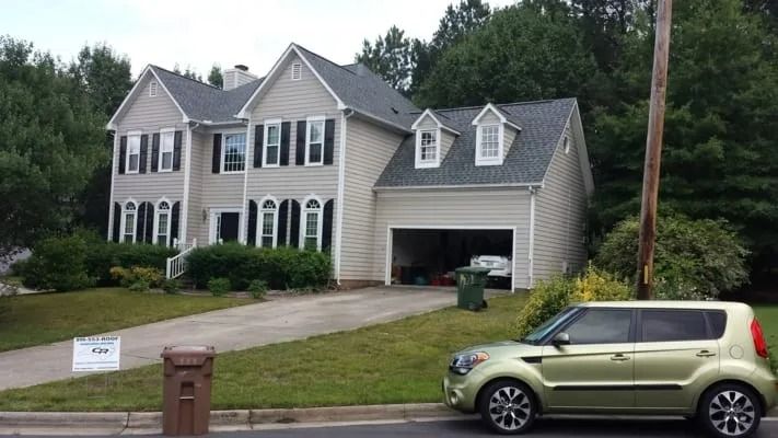 Two-story beige house with a driveway and a green Kia Soul parked in front. Garage door is open, revealing a car inside.