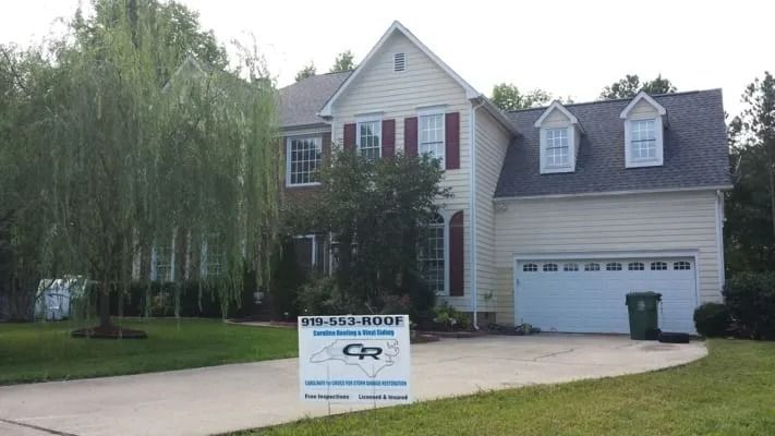 A two-story yellow house with a dark gray roof, a driveway, and a sign advertising roofing services.