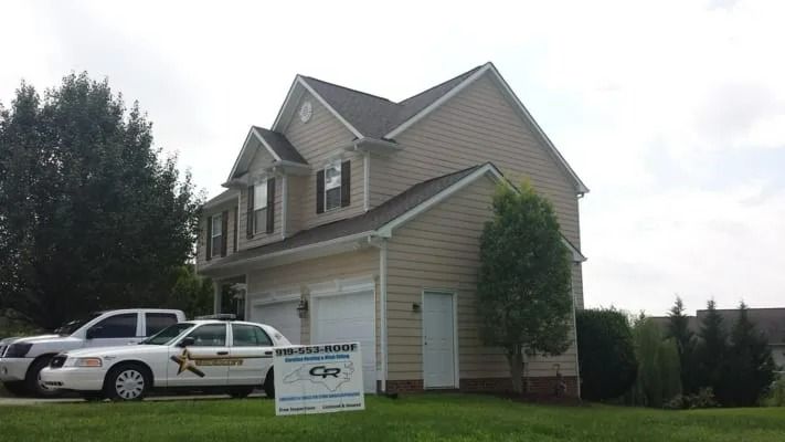 Two-story beige house with a sheriff's vehicle parked in the driveway on a grassy lawn under a cloudy sky.