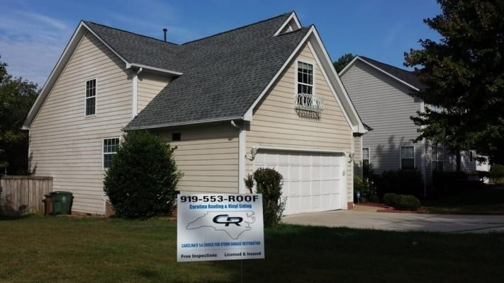 A two-story house with gray roof, light siding, and a garage. A sign in the yard advertises a roofing company.