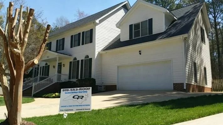 White two-story house with black shutters, garage door, and a sign in the yard.