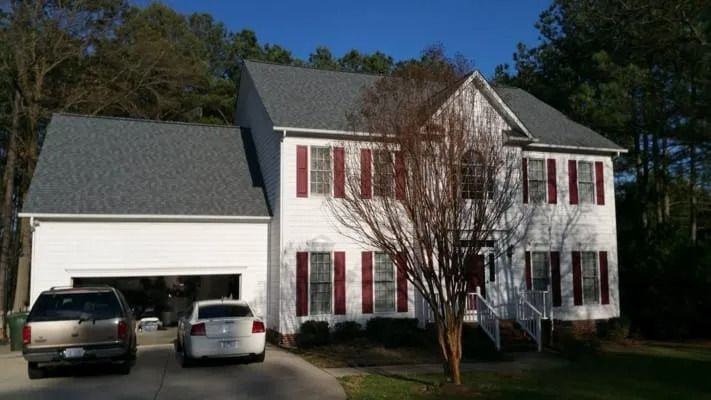 Two-story white house with red shutters, attached garage, and vehicles in the driveway, set amongst trees under a blue sky.