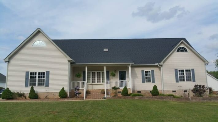 Cream-colored ranch-style home with blue shutters, a dark roof, and a porch on a grassy lawn under a cloudy sky.