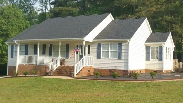 White house with a dark roof, blue shutters, and a porch with an American flag. Set in a grassy yard.