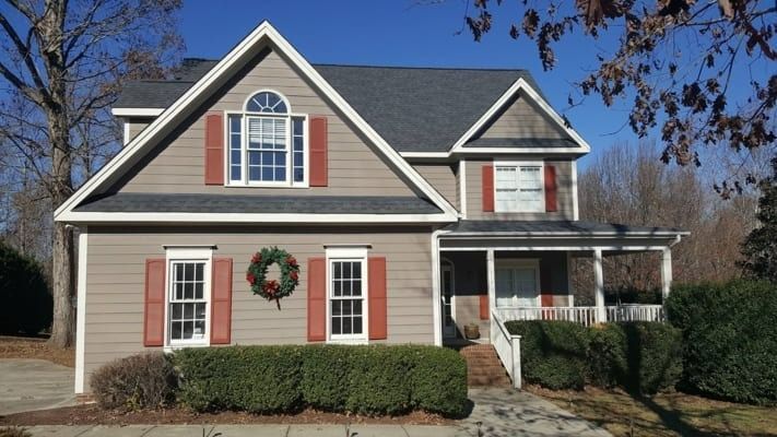 Two-story house with tan siding, red shutters, and a dark gray roof, with a small porch and a green hedge in front.