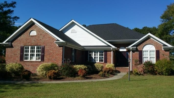 Brick house with dark roof, white trim, and landscaping on a sunny day. Green grass and a clear blue sky.