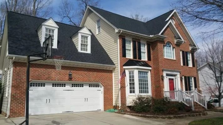 Two-story brick house with a white garage door and a basketball hoop in the driveway. The front door is red, and the roof is black.