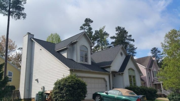 A two-story house with white siding, black roof, and a green convertible in the driveway on a sunny day.