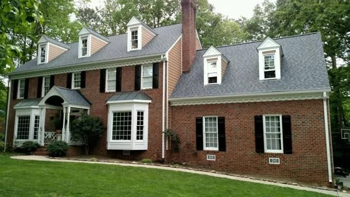 Two-story brick house with black shutters, dormers, and a green lawn.