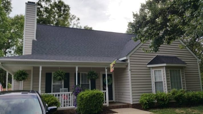Tan house with a dark gray roof, white porch, and black shutters. Green bushes and trees surround the home.