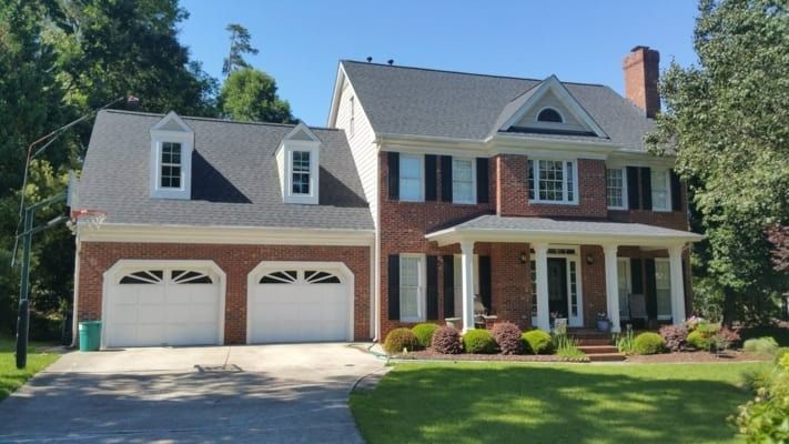 Two-story brick house with white garage doors and black shutters under a clear blue sky. A green lawn and landscaping surround the home.