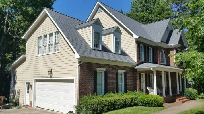 Two-story house with brick and tan siding, gray roof, white garage door, and green bushes.