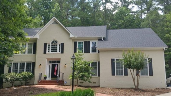 Two-story beige house with black shutters and a dark gray roof, set against a background of green trees.