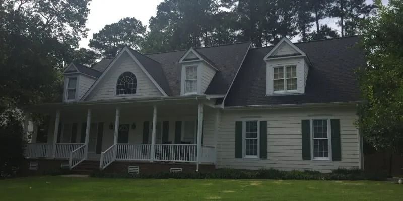 A white house with a large porch and green shutters, surrounded by trees.