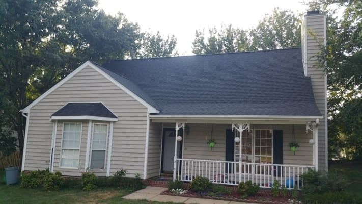 Beige house with black roof, white porch, and chimney, set against a backdrop of trees under a bright sky.