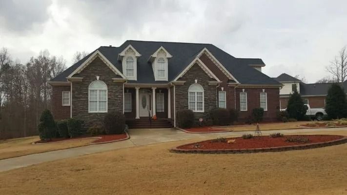 A large brick house with a dark roof and white trim on a cloudy day, with a circular flowerbed and a long driveway.