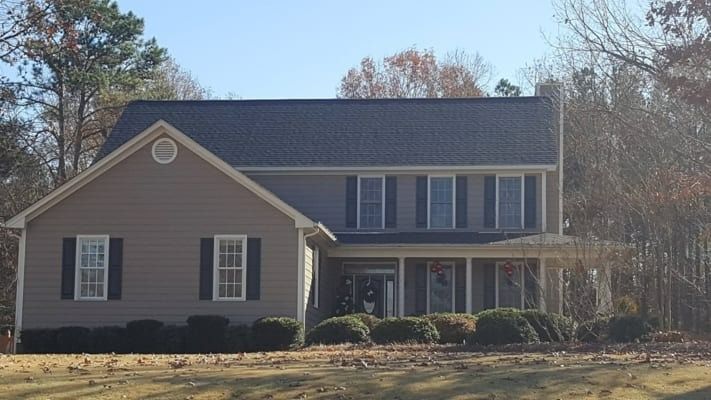 Two-story house with a gray exterior and black shutters; a porch in front. It's surrounded by brown grass and trees.