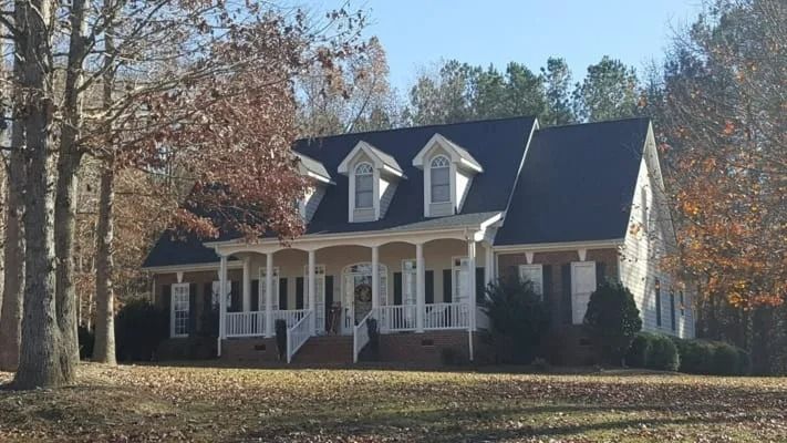 A brick and white colonial house with a long front porch and dormers in a wooded area.