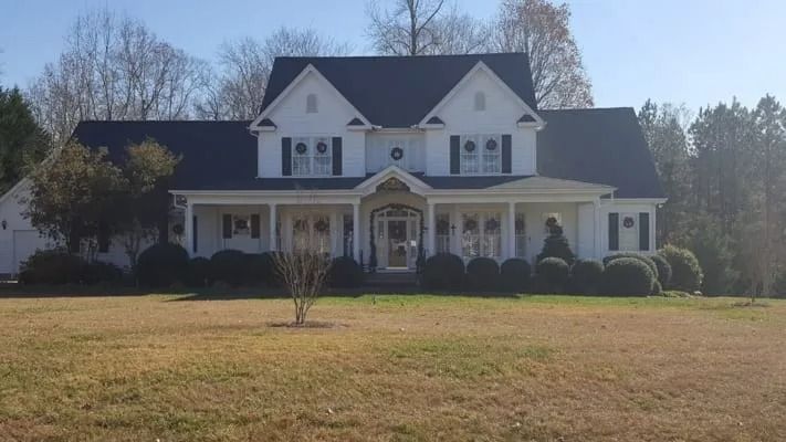 White two-story house with a large porch and dark roof. The house sits on a grassy lawn with trees in the background.