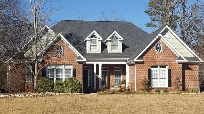 Brick house with a dark gray roof, white trim, and three dormer windows. The house is set against a backdrop of trees and a brown lawn.