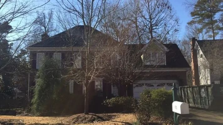 Two-story brick house with white garage door, shutters, and a dormer window. The house is surrounded by bare trees and shrubs.
