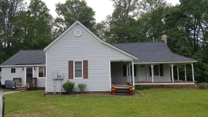 A light gray house with a dark gray roof, a porch, and a small yard with trees in the background.